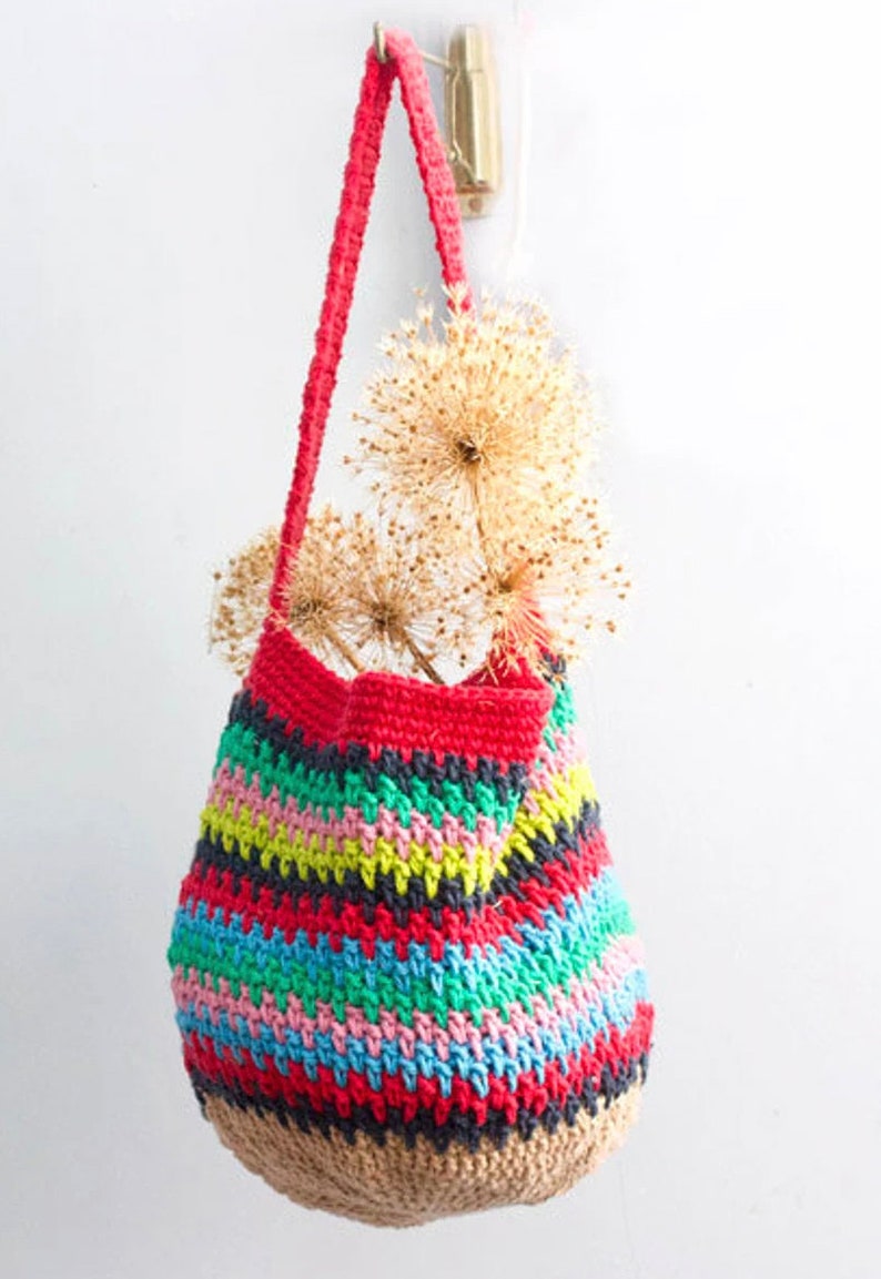 May include: A colorful striped crocheted tote bag with a red handle. The bag is hanging from a hook on a white wall. The bag is filled with dried flowers.