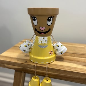 Peut inclure: Une figurine décorative en pots de terre cuite empilés. Le pot supérieur présente un visage peint avec de grands yeux et un nez en forme de cœur rose. Le corps est jaune avec des motifs d'abeilles, et les bras et jambes sont des pots.