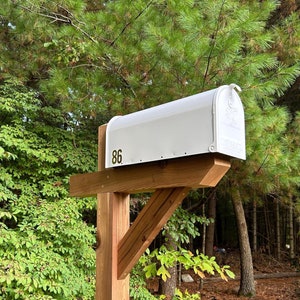 May include: A white mailbox with the number 86 on it is mounted on a wooden post. The post is made of brown wood and has a wooden brace.