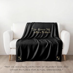 May include: A black blanket draped over a white sofa. The blanket features gold text that reads "Some stories are truly written in the stars." Gold starburst designs are in the corners. The blanket is a home decor item.