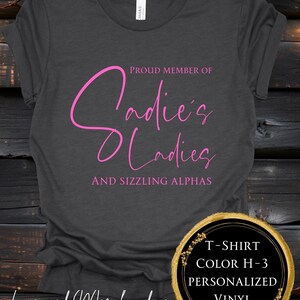 May include: Charcoal grey t-shirt featuring pink text: "Proud Member of Sadie's Ladies and Sizzling Alphas". A circular graphic indicates colour H-3, personalised vinyl, and licensed merchandise.