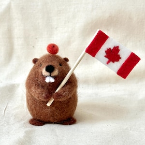 May include: A brown needle-felted beaver figurine with a small red sphere on its head. The beaver has two white felt teeth and is holding a miniature Canadian flag made of white and red felt attached to a thin wooden stick in its front paws.