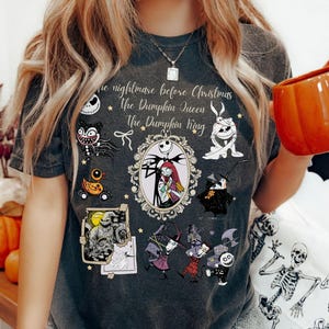 Pu&ograve; includere: T-shirt grigio scuro con personaggi e testo di "Nightmare Before Christmas". Il design include Jack Skellington e Sally in una cornice ovale, insieme ad altri personaggi e le frasi "The Pumpkin Queen" e "The Pumpkin King".