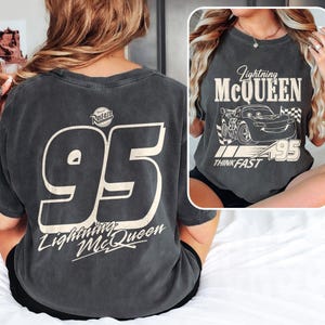 May include: Dark gray t-shirt featuring the number 95 in a large, off-white font on the back, with "Lightning McQueen" below. The front of the shirt has a graphic of Lightning McQueen with the text "Lightning McQueen", "Think Fast", and the number 95.