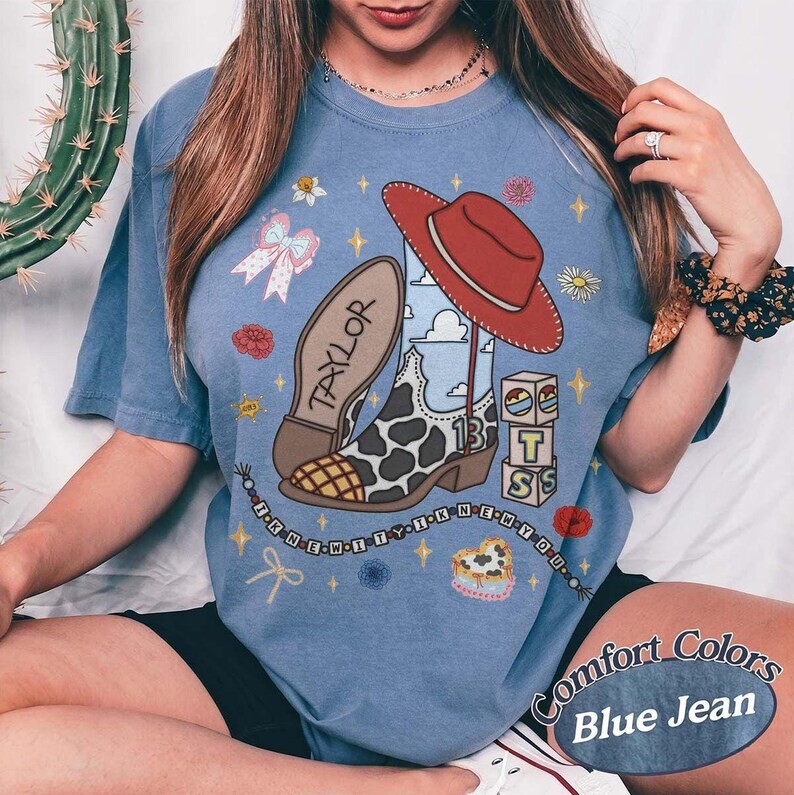 May include: A blue jean colored t-shirt featuring a graphic print of a cow print cowboy boot, a red cowboy hat, and blocks with the letters T, S, and the number 13. The design includes the text Taylor and I KNEW IT I KNEW YOU.