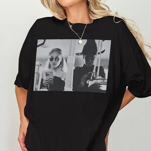 May include: Black oversized t-shirt featuring a black and white image of two people. One person is wearing sunglasses and holding a phone, the other is wearing a hat. The shirt has a round neckline and short sleeves.