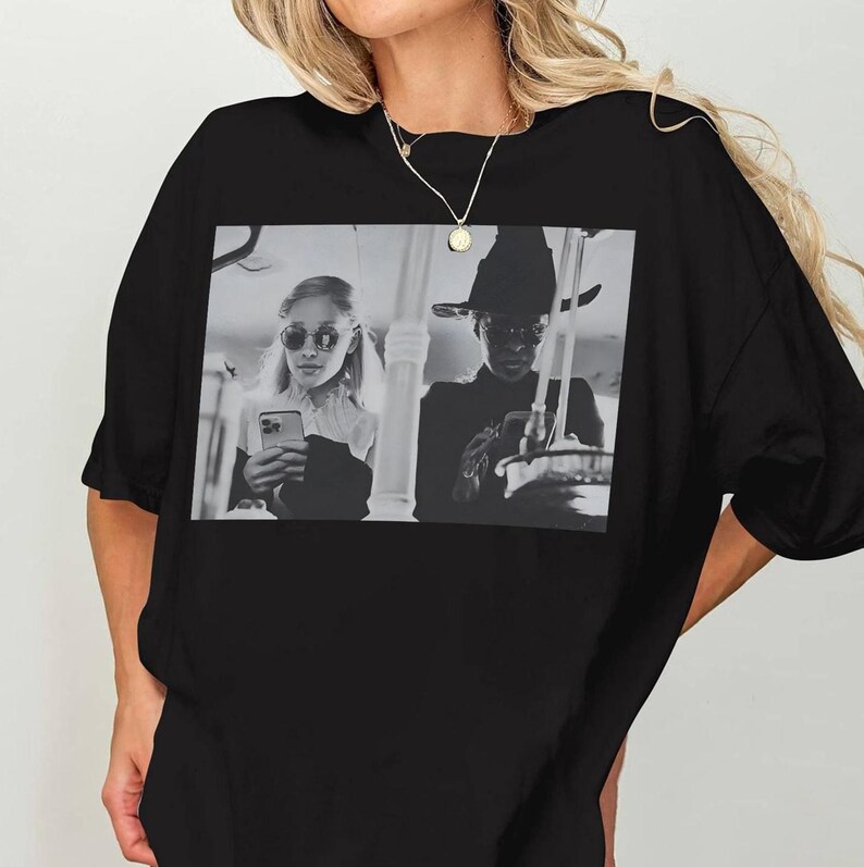 May include: Black oversized t-shirt featuring a black and white image of two people. One person is wearing sunglasses and holding a phone, the other is wearing a hat. The shirt has a round neckline and short sleeves.