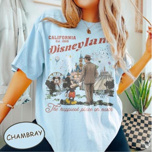 Puede incluir: Camiseta azul claro con un gr&aacute;fico de Disneyland. El gr&aacute;fico incluye a Mickey Mouse, un hombre con traje, una noria y un castillo. El texto en la camiseta dice "CALIFORNIA Est. 1955 Disneyland" y "The happiest place on earth."