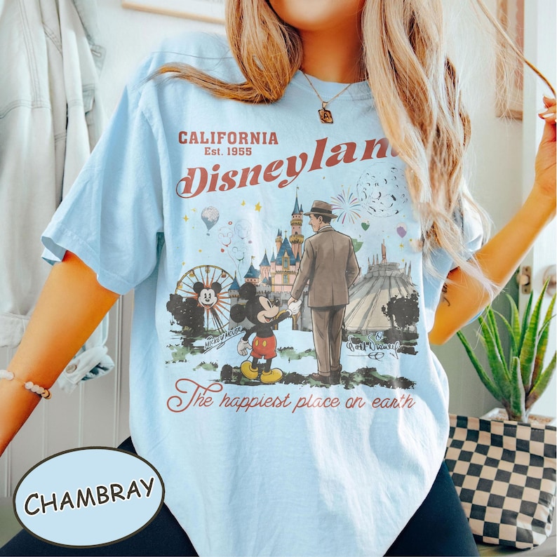 Puede incluir: Camiseta azul claro con un gr&aacute;fico de Disneyland. El gr&aacute;fico incluye a Mickey Mouse, un hombre con traje, una noria y un castillo. El texto en la camiseta dice "CALIFORNIA Est. 1955 Disneyland" y "The happiest place on earth."