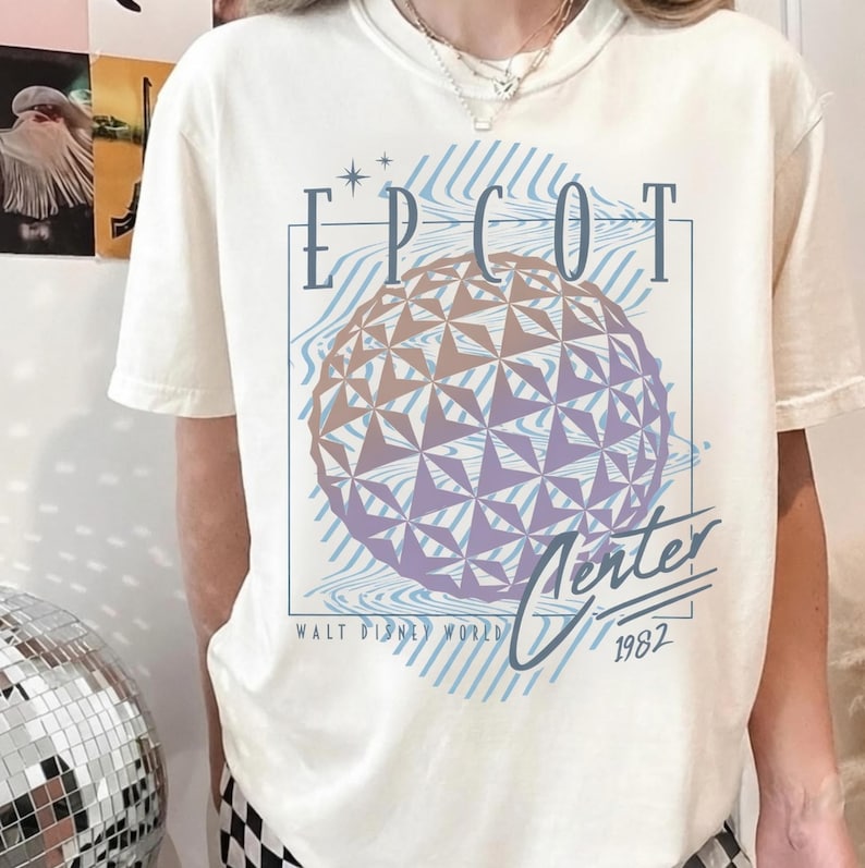 May include: Cream-colored t-shirt featuring a graphic of the EPCOT Center sphere with text that reads "EPCOT Center" and "Walt Disney World 1982". The design includes blue and purple accents.