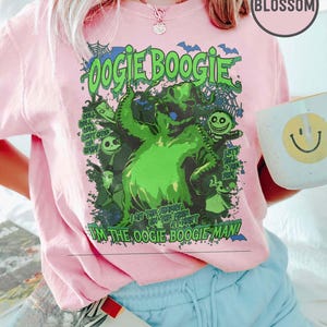 Op de afbeelding: Roze T-shirt met een groene afbeelding van Oogie Boogie uit The Nightmare Before Christmas. Het ontwerp bevat de tekst "Oogie Boogie" en "I'm the Oogie Boogie Man!" Een witte mok met een smiley is zichtbaar.