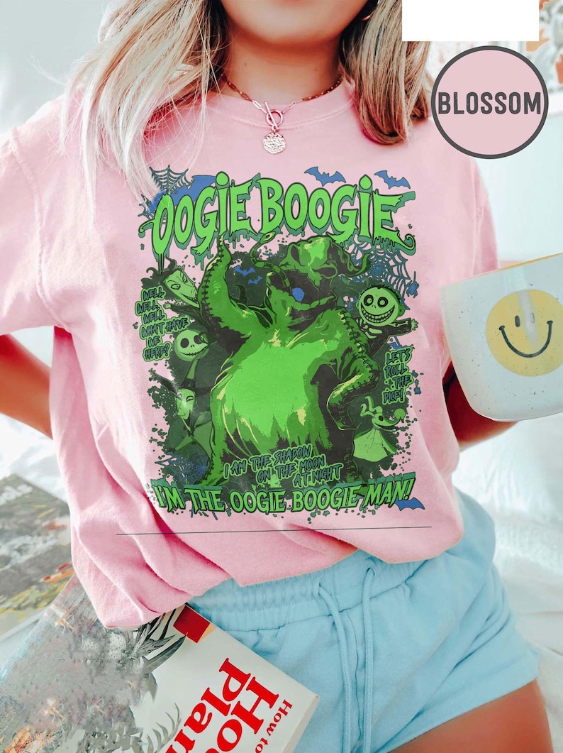 Op de afbeelding: Roze T-shirt met een groene afbeelding van Oogie Boogie uit The Nightmare Before Christmas. Het ontwerp bevat de tekst "Oogie Boogie" en "I'm the Oogie Boogie Man!" Een witte mok met een smiley is zichtbaar.