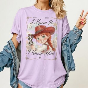 Puede incluir: Una camiseta de color morado claro con un gr&aacute;fico de una vaquera de dibujos animados con un sombrero marr&oacute;n y un guante con estampado de vaca. El texto dice "EST 2026", "I Knew It" y "I Knew You" con una nota musical. Borde de cuerda.