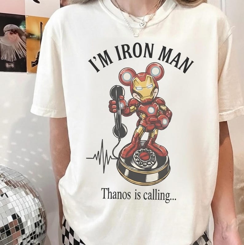 Op de afbeelding: Cr&egrave;mekleurig T-shirt met een afbeelding van Iron Man als muis, die een telefoon vasthoudt. De tekst "I'M IRON MAN" staat boven de afbeelding en "Thanos is calling..." eronder. Een hartslaglijn staat links.