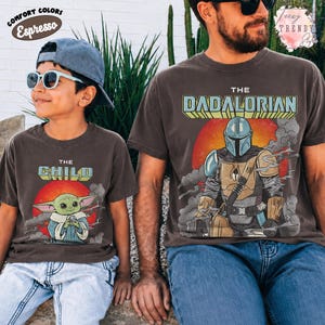 May include: Two dark gray t-shirts featuring graphic prints. The smaller shirt displays a green creature with large ears and the text THE CHILD. The larger shirt shows a masked character in armor with the text THE DADALORIAN over a red sun.