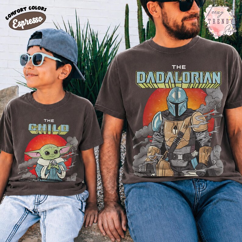 May include: Two dark gray t-shirts featuring graphic prints. The smaller shirt displays a green creature with large ears and the text THE CHILD. The larger shirt shows a masked character in armor with the text THE DADALORIAN over a red sun.