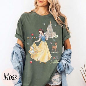 K&ouml;nnte beinhalten: Moosgr&uuml;nes T-Shirt mit einer Aquarellillustration von Schneewittchen in einem gelben Kleid, mit einem Schloss, einem Reh und V&ouml;geln. Der Text "Moss" befindet sich in einem rosa Kreis unten links.