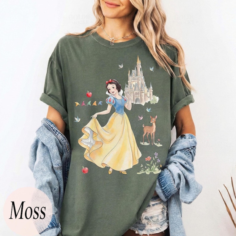 K&ouml;nnte beinhalten: Moosgr&uuml;nes T-Shirt mit einer Aquarellillustration von Schneewittchen in einem gelben Kleid, mit einem Schloss, einem Reh und V&ouml;geln. Der Text "Moss" befindet sich in einem rosa Kreis unten links.