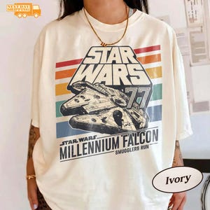 Può includere: T-shirt color avorio con un design di Star Wars. La grafica include il testo "STAR WARS" sopra un'immagine del Millennium Falcon, con "77" e "MILLENNIUM FALCON SMUGGLERS RUN" sotto. Il design ha uno schema di colori retrò.