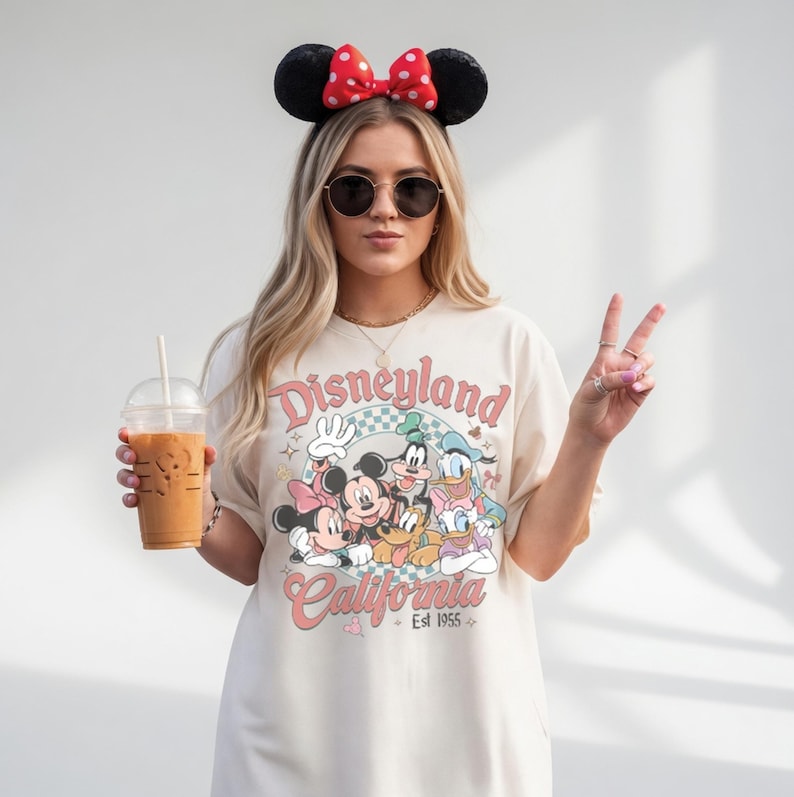May include: Cream-colored t-shirt with a Disneyland California graphic featuring Mickey Mouse, Minnie Mouse, Donald Duck, and Goofy. The shirt has a vintage aesthetic. The person is wearing a Minnie Mouse ear headband and sunglasses.