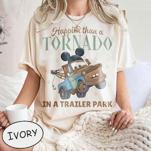 Può includere: T-shirt color avorio con un design di carro attrezzi dei cartoni animati, che incorpora le orecchie di Topolino, e la scritta "Happier than a Tornado in a Trailer Park". Il design presenta colori marrone, blu e verde. La maglietta è indossata da una persona.