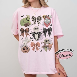 May include: A light pink t-shirt featuring a graphic print of characters and bows from The Nightmare Before Christmas, including Jack Skellington, Sally, Oogie Boogie, Zero, and various bows, with a Comfort Colors Blossom label in the corner.