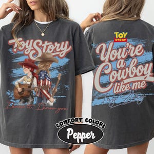 May include: A charcoal grey t-shirt featuring a Toy Story graphic. The front displays two figures in cowboy hats with guitars and the text "Toy Story" and "I know it, I know you". The back features the text "Toy Story" and "You're a Cowboy like me".