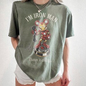 Op de afbeelding: Olijfgroen T-shirt met een Iron Man-stripfiguur met Mickey Mouse-oren die een telefoon vasthoudt. De tekst "I'M IRON MAN" staat boven het personage en "Thanos is calling..." eronder. Er is ook een hartslaggrafiek aanwezig.