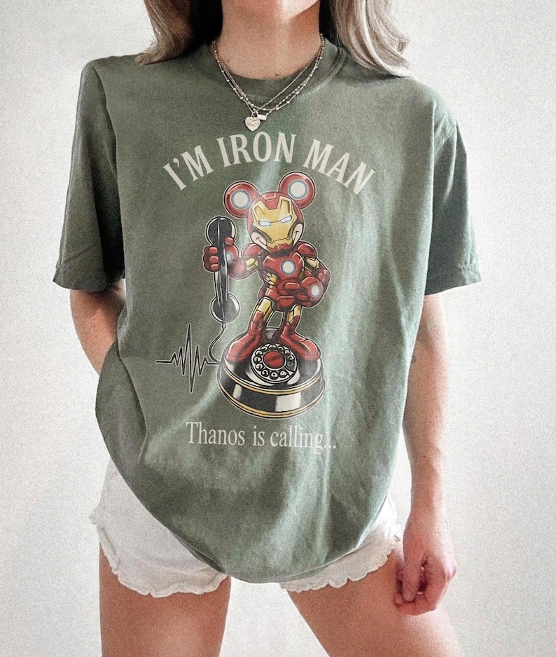 Op de afbeelding: Olijfgroen T-shirt met een Iron Man-stripfiguur met Mickey Mouse-oren die een telefoon vasthoudt. De tekst "I'M IRON MAN" staat boven het personage en "Thanos is calling..." eronder. Er is ook een hartslaggrafiek aanwezig.