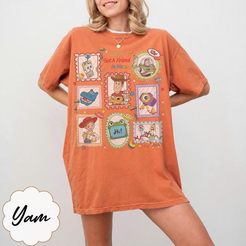 May include: An orange short-sleeve t-shirt featuring a graphic print of various cartoon characters in postage stamp-style frames. The central text reads "You've Got A Friend In Me" and "Hi!" on a green frog-shaped frame. The design includes multiple colourful figures.