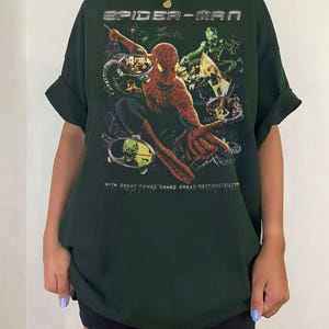 May include: Dark green Spider-Man t-shirt featuring a graphic of Spider-Man and other characters. The text "SPIDER-MAN" is at the top, and "WITH GREAT POWER COMES GREAT RESPONSIBILITY" is at the bottom.