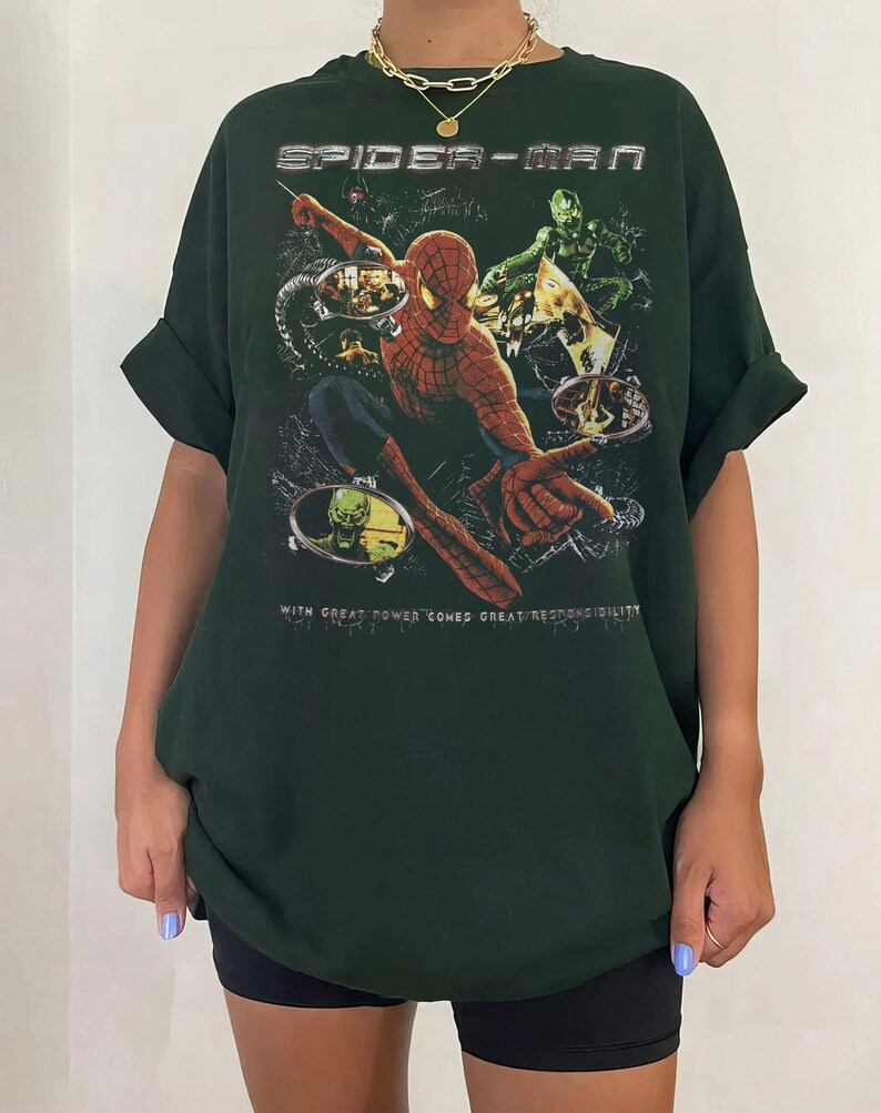 May include: Dark green Spider-Man t-shirt featuring a graphic of Spider-Man and other characters. The text "SPIDER-MAN" is at the top, and "WITH GREAT POWER COMES GREAT RESPONSIBILITY" is at the bottom.