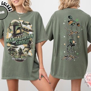 Pode incluir: Uma t-shirt verde musgo com uma impress&atilde;o gr&aacute;fica de The Mandalorian e Grogu na frente com o texto "The Mandalorian and Grogu" e "This is the way". A parte de tr&aacute;s da t-shirt exibe um alinhamento vertical de quatro ve&iacute;culos Star Wars.