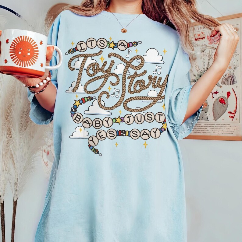 May include: A light blue t-shirt featuring a graphic print with the text "ITS A Toy Story BABY JUST YES SAY" written in a rope-style font. The design includes colorful beads, small stars, musical notes, and white clouds on a light blue background.