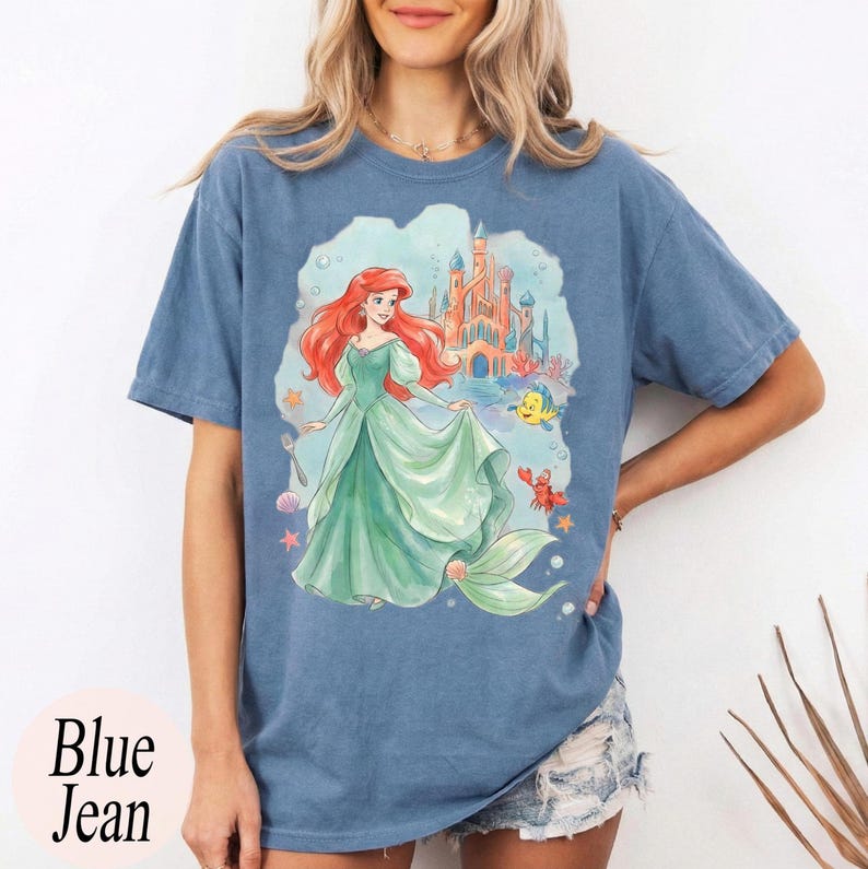 K&ouml;nnte beinhalten: Blaues Jeans-T-Shirt mit einer Aquarellillustration einer Meerjungfrau in einem gr&uuml;nen Kleid, mit roten Haaren, einem Schloss und Meeresbewohnern. Auf dem Shirt steht "Blue Jean" unten links.