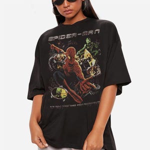May include: Black t-shirt featuring a Spider-Man graphic. The design includes Spider-Man in action, the Green Goblin, and the text "SPIDER-MAN" and "WITH GREAT POWER COMES GREAT RESPONSIBILITY". The shirt has short sleeves and a relaxed fit.