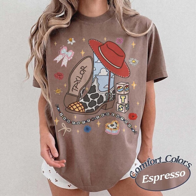 May include: An espresso brown t-shirt featuring a graphic of a cowboy boot with a cow print pattern, a red hat, and the name TAYLOR. The design includes blocks with the letters T, S, and the number 13, plus a beaded string with the text I KNEW YOU.