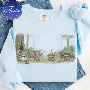 K&ouml;nnte beinhalten: Ein hellblaues Sweatshirt mit Rundhalsausschnitt und einer grafischen Illustration von vier kleinen gr&uuml;nen Wesen in einer Waldumgebung. Der Druck zeigt die Figuren beim Interagieren mit einer schwebenden Metallkugel, einer Tasse und einer Kapsel, mit dem Text coo.