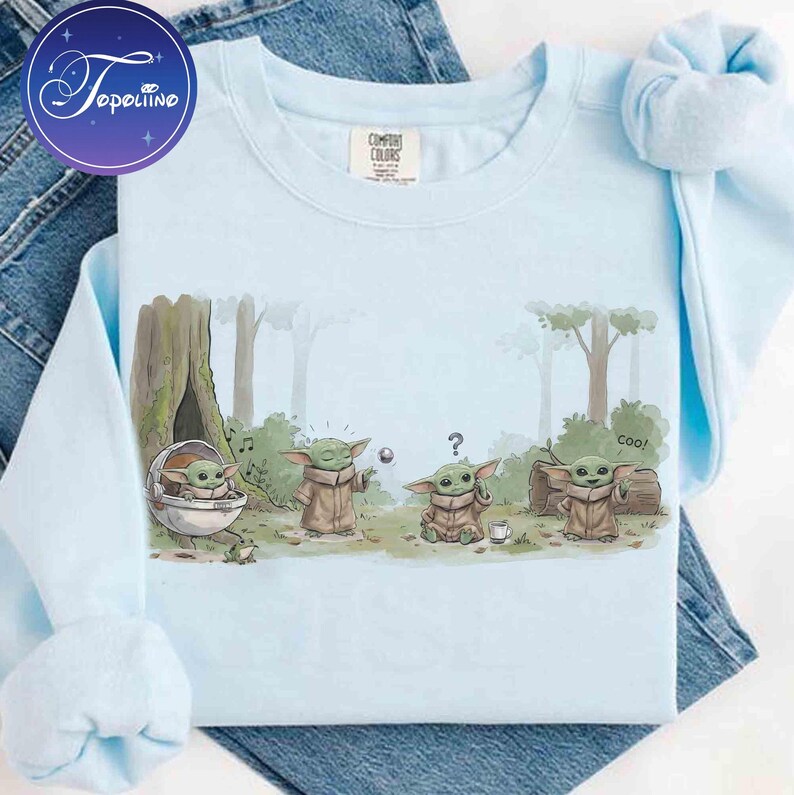 K&ouml;nnte beinhalten: Ein hellblaues Sweatshirt mit Rundhalsausschnitt und einer grafischen Illustration von vier kleinen gr&uuml;nen Wesen in einer Waldumgebung. Der Druck zeigt die Figuren beim Interagieren mit einer schwebenden Metallkugel, einer Tasse und einer Kapsel, mit dem Text coo.