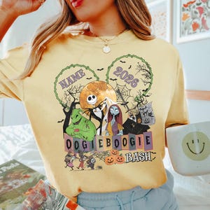 May include: A pale yellow t-shirt featuring a graphic print of characters from The Nightmare Before Christmas, including Jack Skellington, Sally, and Oogie Boogie. The design includes the text NAME, 2026, and OOGIE BOOGIE BASH with pumpkins.
