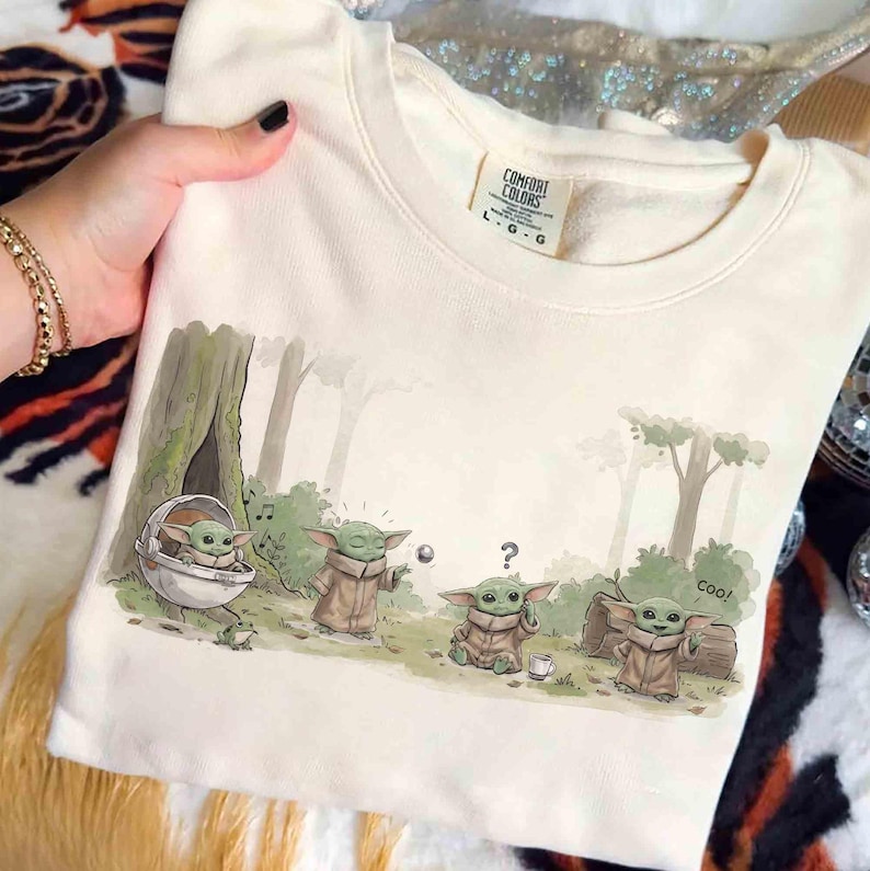 K&ouml;nnte beinhalten: Ein cremefarbenes Sweatshirt mit Rundhalsausschnitt und einer grafischen Illustration von vier kleinen gr&uuml;nen Wesen im Wald. Der Druck zeigt eines in einer schwebenden Kapsel, eines nutzt die Macht, eines wirkt verwirrt und eines winkt mit Text coo!