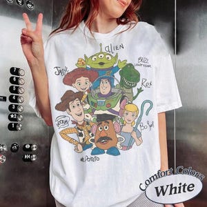 May include: A white t-shirt featuring a graphic print of Toy Story characters including Woody, Buzz Lightyear, Jessie, Rex, an alien, Mr. Potato Head, and Bo Peep. The names of each character are handwritten in black ink next to their respective figures.