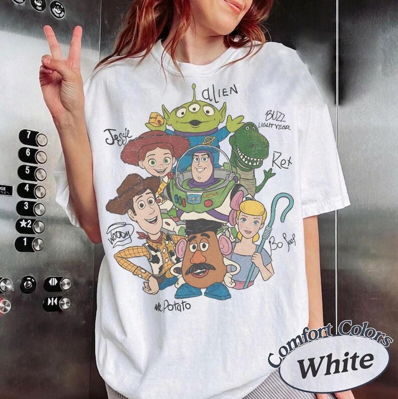 May include: A white t-shirt featuring a graphic print of Toy Story characters including Woody, Buzz Lightyear, Jessie, Rex, an alien, Mr. Potato Head, and Bo Peep. The names of each character are handwritten in black ink next to their respective figures.