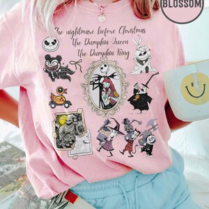 Pu&ograve; includere: T-shirt rosa chiaro con personaggi e testo di "Nightmare Before Christmas". Il design include Jack Skellington, Sally e altri personaggi. Il testo recita "The nightmare before Christmas, the Pumpkin Queen, The Pumpkin King."