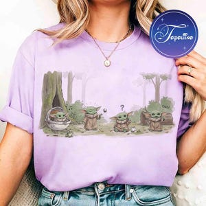 K&ouml;nnte beinhalten: Ein hellviolettes T-Shirt mit einer grafischen Illustration von vier kleinen gr&uuml;nen Wesen in einer Waldumgebung. Ein Wesen sitzt in einer Kapsel, eines greift nach einer schwebenden Kugel, eines wirkt verwirrt mit einem Fragezeichen und eines winkt mit dem Text coo.