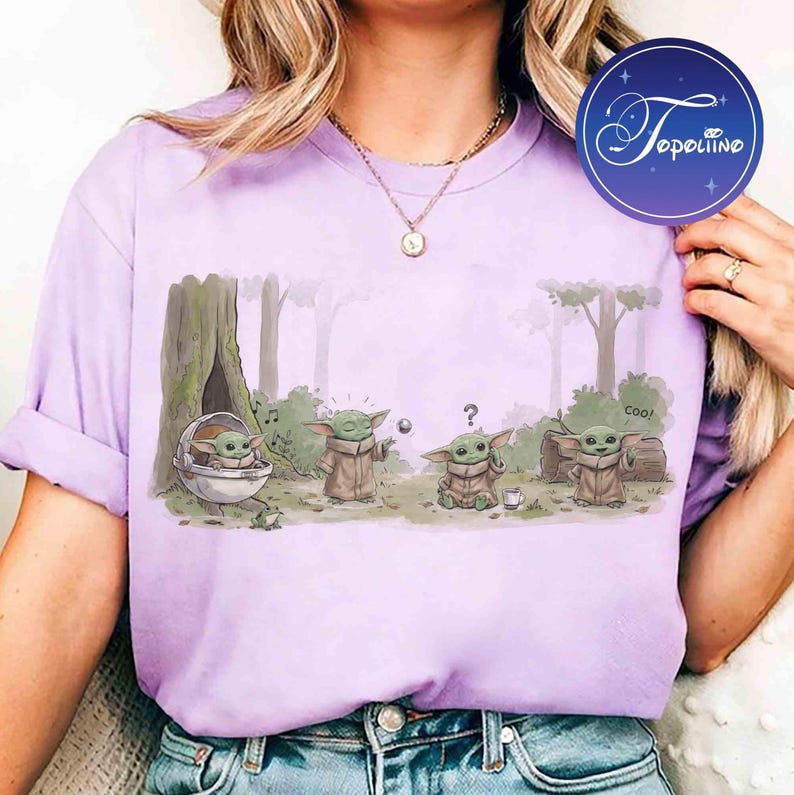 K&ouml;nnte beinhalten: Ein hellviolettes T-Shirt mit einer grafischen Illustration von vier kleinen gr&uuml;nen Wesen in einer Waldumgebung. Ein Wesen sitzt in einer Kapsel, eines greift nach einer schwebenden Kugel, eines wirkt verwirrt mit einem Fragezeichen und eines winkt mit dem Text coo.