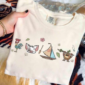 Pode incluir: Uma camisola de gola redonda cor de creme com etiqueta Comfort Colors e uma impress&atilde;o gr&aacute;fica de um galo, um leit&atilde;o, um remo de madeira, um veleiro e uma personagem de coco com uma lan&ccedil;a, rodeados por pequenas flores tropicais e folhas.