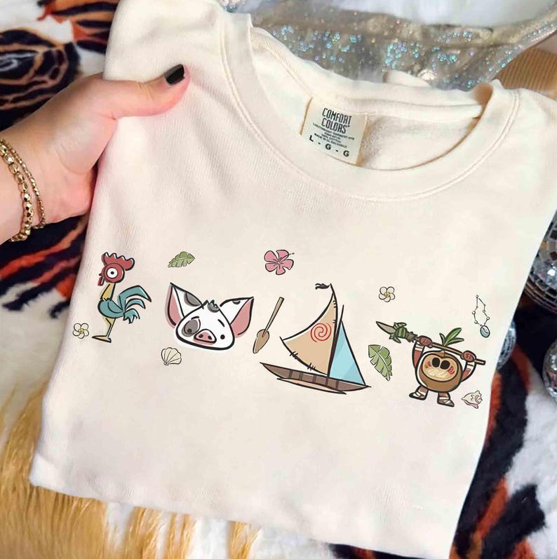 Pode incluir: Uma camisola de gola redonda cor de creme com etiqueta Comfort Colors e uma impress&atilde;o gr&aacute;fica de um galo, um leit&atilde;o, um remo de madeira, um veleiro e uma personagem de coco com uma lan&ccedil;a, rodeados por pequenas flores tropicais e folhas.