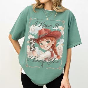 Puede incluir: Una camiseta de manga corta color verde salvia con el gr&aacute;fico de una vaquera de dibujos animados con pelo rojo y sombrero marr&oacute;n. El texto dice "EST 2026", "I Knew It" y "I Knew You" rodeado por un borde de cuerda, estrellas y herraduras.
