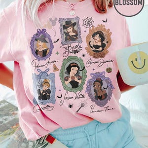 May include: Pink t-shirt featuring framed portraits of Disney princesses, including Belle, Aurora, Jasmine, Snow White, Cinderella, and Tiana. The shirt has a round "Blossom" logo and handwritten princess names.